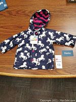 Girls raincoat laid flat on wooden surface showing unicorn print, pink-striped hood lining, front pockets, and buttons.