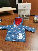 Blue boys raincoat with white tractor pattern and red fleece lining inside. Laid flat on a wooden table with Hatley branded shopping bag behind it.
