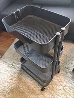 Front angled view of gray Ikea RASKOG utility cart showing three mesh trays and caster wheels on light carpet.