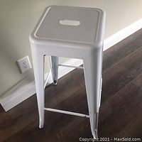 White iron bar stool 76 cm height with square seat, central carry handle, footrests on all sides, standing on wooden floor near wall socket.