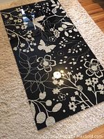 Rectangular glass table top laid flat on a light surface showing clear etched floral and butterfly pattern on black background.