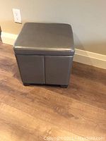 Grey square ottoman with closed top, upholstered in smooth grey faux leather or vinyl, standing on a wood floor.