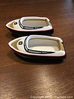 Two ceramic nautical boats shown on wooden table. One boat shown assembled, demonstrating the cavity to hold dip or spread, painted white interior. Exterior painted to resemble a small boat with red, beige, and black details.