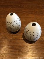 Pair of small, white ceramic, egg-shaped flower holders with gold dots, shown on a wooden surface.