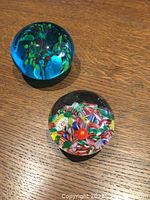 Both vintage glass paperweights on a wooden surface showing one blue floral design and one colorful millefiori cluster design