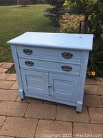 Front view of vintage light blue wooden chest showing two drawers, decorative metal handles, and double cupboard doors, placed on outdoor paved surface with grass background.