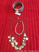 Top view showing all three jewelry pieces on red fabric background: long necklace with dangling silver discs, rhinestone bead bracelet, taupe leather braided bracelet.