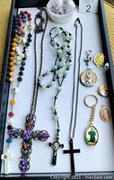 Full view of religious jewelry lot on white tray showing six necklaces, several pendants, a keychain, and rosary beads.