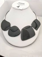 Front view of avant-garde style black asymmetrical necklace on white display bust showing five geometric pieces with stitched designs on black cord.