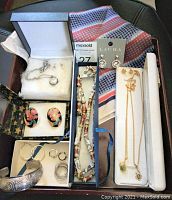 Wood tray containing various costume jewelry including necklaces, earrings, rings, and a retro scarf in background.