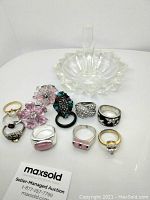 Image of glass ring holder and 9 assorted fashion rings arranged around it on a white background.