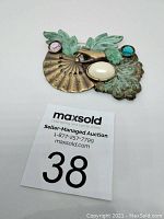 Brooch overview showing green patina leaves, brass fan, pearl centerpiece, and colored stones photographed with auction card.