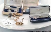 Overview of assorted men's cufflinks, tie clips, and lapel pins arranged on a white surface, showing original boxes and varied designs.