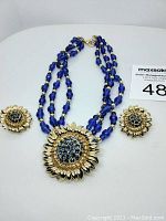 Front view of the necklace and earrings laid flat on a white surface showing the blue beads, gold-tone sunflower pendant with blue stones, and matching earrings.