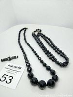 Two black glass beaded necklaces and one black lace and metal lapel pin on a white surface alongside auction lot number card.