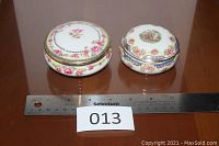 Two small round porcelain dresser boxes with lids on wooden surface, with ruler for scale, one marked KPM with roses, the other unmarked with people scene.
