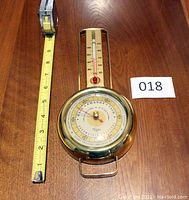 Photo showing the front view of the Taylor hanging wall barometer and thermometer next to a measuring tape indicating length about 10.25 inches.