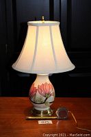 Front view of the Moorcroft lamp with white shade and floral decoration on the bulbous cream base illuminated from within.