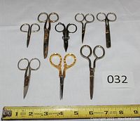 Eight small scissors arranged on white surface with measuring tape showing length between 2.75 and 5.25 inches, all metal with different handle styles including one pair with gold decorative handles.