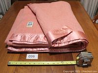 Photo showing folded rose wool blanket with satin binding and measuring tape for scale.