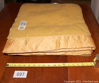 Folded gold-coloured wool blanket with a measuring tape showing dimensions and a label with the lot number in view.
