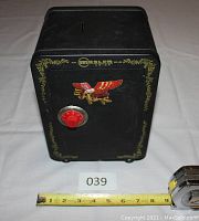 Front view of black Mosler Jr. bank safe with coin slot on top, red combination dial and eagle decal on door, with measuring tape for size reference.