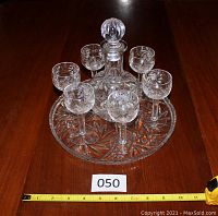 Full view of crystal pinwheel liquor decanter with six matching glasses arranged on round serving plate placed on wood surface. Tape measure shows plate diameter about 10 inches.