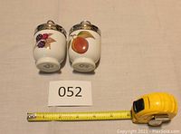 Pair of Royal Worcester egg coddlers side by side with tape measure showing dimensions approximately 3 3/8 inches tall and 2 1/8 inches diameter, each featuring fruit designs and chrome lids with loop handles.