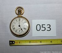 Front view showing white dial with black Roman numerals; sub-dial at 6 o'clock; gold-tone crown and bow at top; measuring tape showing approx. 2 inch diameter.