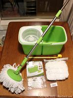 Green and white Big Boss InstaMop bucket with dual compartments and spinning wringer, mop with stainless steel handle and green connectors, bonus mop head sealed in plastic, manual and packaging on wooden table.