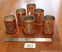 Front view of all six copper Moscow mule mugs arranged in two rows with a ruler and identification card in foreground to show scale.