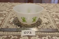 Front view of white opaque mixing bowl with lime green clown designs on a lace tablecloth beside a steel ruler.