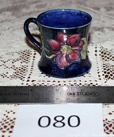 Front side view showing burgundy/red clematis flower with green foliage on deep cobalt blue background, small ceramic tankard with handle and ruler for scale.