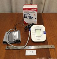 Photo showing Omron blood pressure monitor front and cuff with original box in background and ruler for scale.