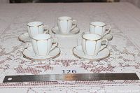 Five white porcelain demitasse cups with matching saucers arranged in a group on a lace tablecloth. Each cup and saucer features gold hand painted accents showing minor wear.
