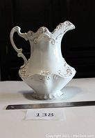 Front side view of a white ceramic Victorian-style pitcher with embossed relief and hand-painted gold swirls, 8 inches tall, on white surface with measuring ruler and lot number.