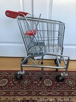 Side view of small metal kids shopping cart showing metal wire basket, red plastic handle, and black plastic wheels on patterned rug.