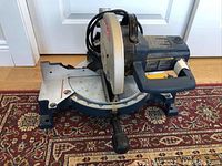 Blue and silver Ryobi mitre saw on carpeted surface with visible angle adjustment and handle.
