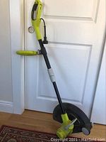 Full view of the Ryobi electric weed wacker leaning against a door, showing the entire tool including handle, shaft, and cutting head with protective shield.