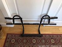 Front view of black metal chin up bar with padded grips and curved handle options, placed on wooden floor against white doors.