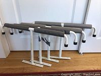 Four white metal T-racks with gray foam padding stacked horizontally, showing front and side angles on wooden floor.