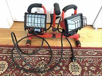 Front view of two red portable flood lights connected on a frame with black handles, power cords visible, wire guards over bulbs