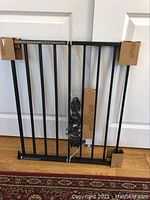Photo of assembled black metal gate frame with vertical bars, unassembled and secured with cardboard and plastic in front of white double doors on wood floor