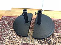 Two black patio umbrella stands, half-circle shaped, with pole holders and adjustable knobs on a patterned carpet.