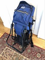 Another angle showing the front and side of the baby backpack carrier upright on a patterned carpet with metal frame.