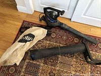 Full set laid out on patterned carpet, showing the main blower unit, collection bag, and mulcher attachment tube.