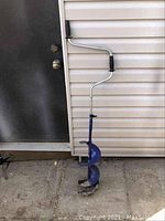 Blue manual post hole digger auger leaning against a beige siding wall and next to a door. The auger has a metal spiral blade and a crank handle with black rubber grips.