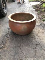 Large round clay pot sitting on an asphalt surface, showing the side and top view of the pot with visible wear and hairline cracks.