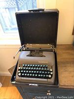 Front view of the vintage Smith Corona typewriter inside its open hard carrying case on a wooden table near a window.