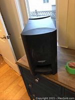 Front view of the black Bose subwoofer speaker with Bose logo on the grille, placed on a wooden surface near window.